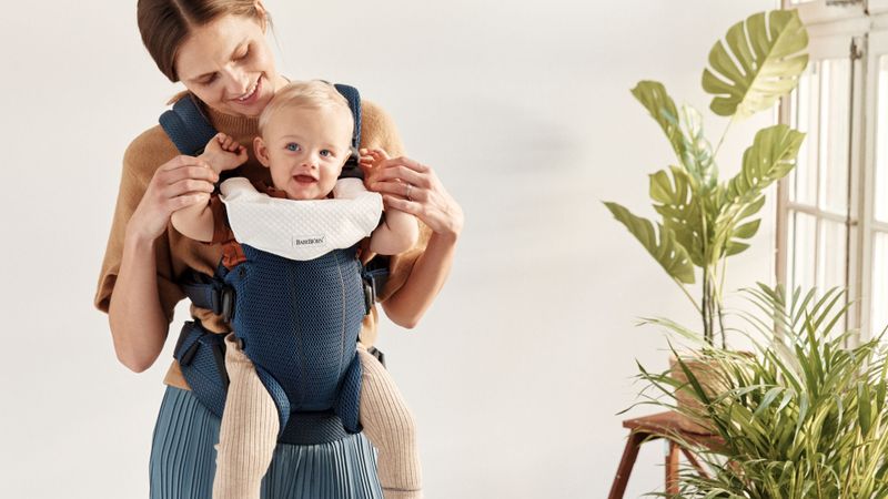 Smiling woman holds a happy baby in a blue carrier with white padding, standing next to green plants.