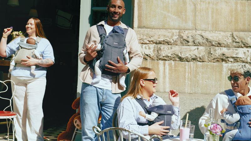 People with baby carriers enjoy a sunny day at an outdoor café, sitting and standing around tables with drinks.