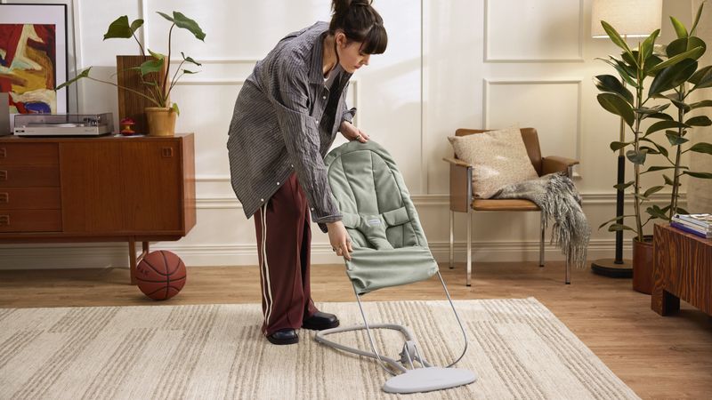 A person adjusts a baby bouncer chair in a cozy living room with a basketball, plants, and a wooden sideboard.
