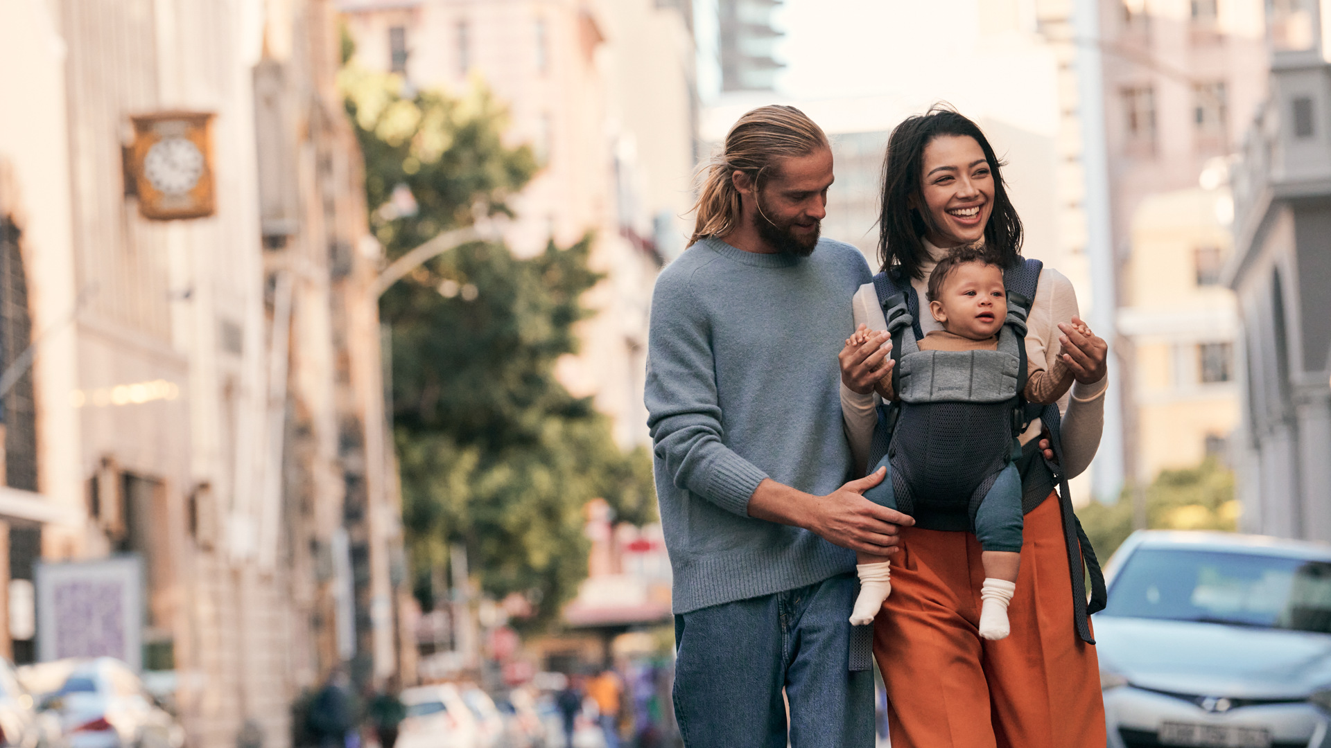 A smiling couple walks down a city street with a baby in a Baby Carrier Harmony. The man wears a gray sweater; the woman wears orange trousers.
