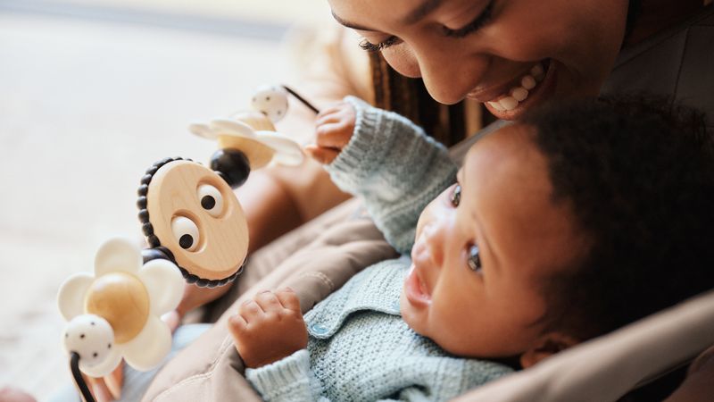 Mother smiling at her baby in a car seat, who is reaching for a hanging toy with a wooden bee and flowers.