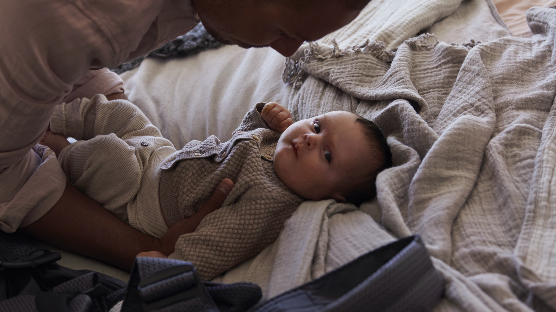 A baby lying on a bed gazes upwards while being gently held by an adult, surrounded by soft blankets and natural light.