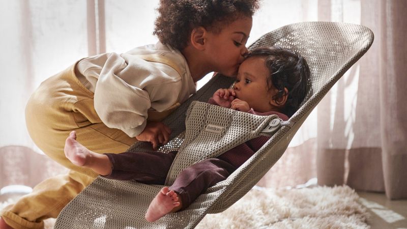 A young child gently kisses a baby sitting in a mesh baby bouncer. They are indoors on a soft rug with light streaming through curtains.