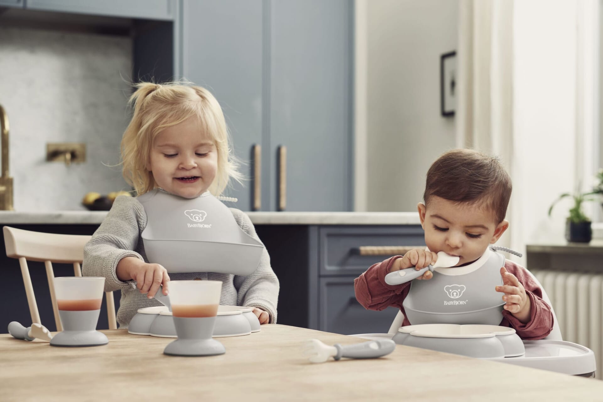 BabyBjörn Kitchen products Two toddlers in bibs eating at a table with bowls and cups. One is smiling while the other is focused on using a spoon. Kitchen setting.