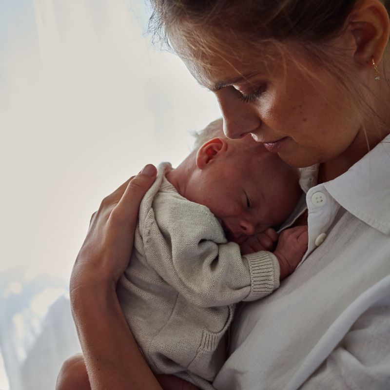 A woman gently cradles a sleeping baby against her chest, both wearing light-colored clothing, with soft natural light in the background.