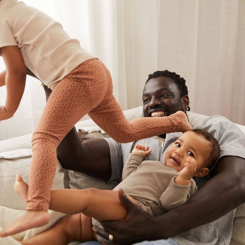 A smiling man holds a baby, while another child playfully climbs over them on a couch. The scene is joyful and cozy.