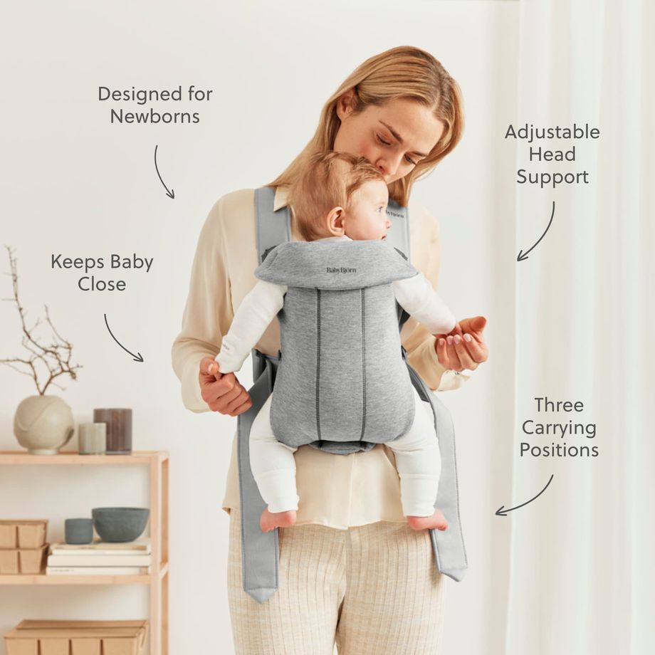 Mother wearing a gray front baby carrier holding a newborn upright against her chest; on-image labels note adjustable head support, newborn-safe design and three positions.