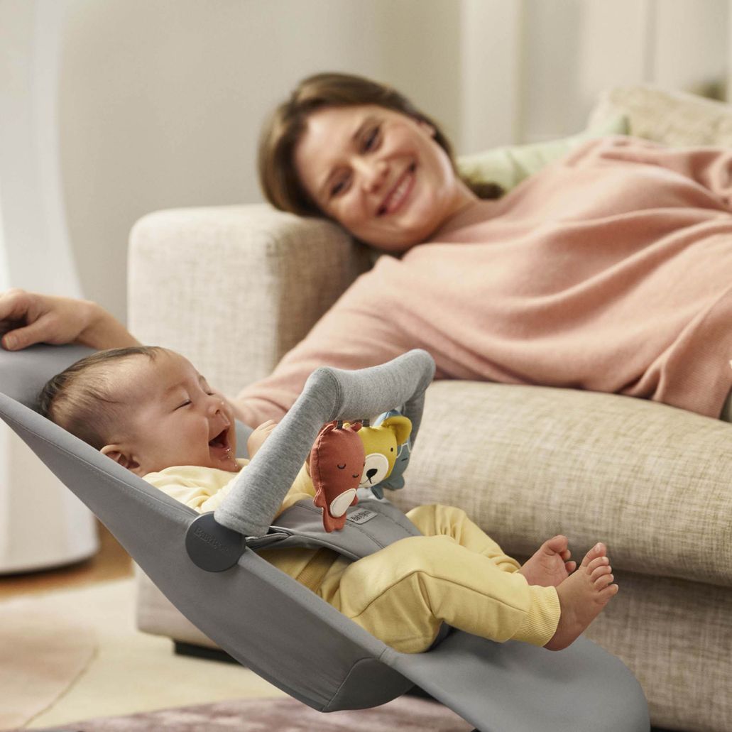 A baby laughs while sitting in a bouncer with toys, as a woman smiles and watches from a sofa in the background.