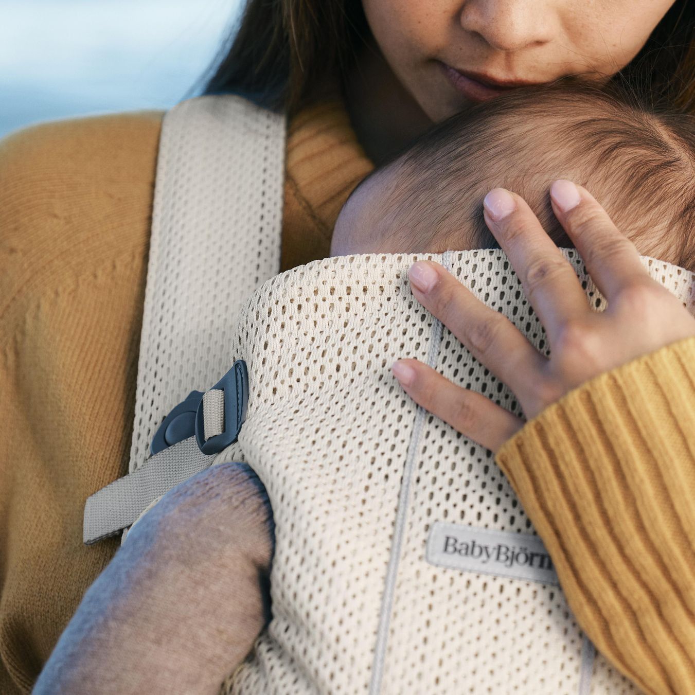 A woman gently cradles a baby in a beige Baby Björn carrier. The baby's head is tenderly held close by the woman's hand.