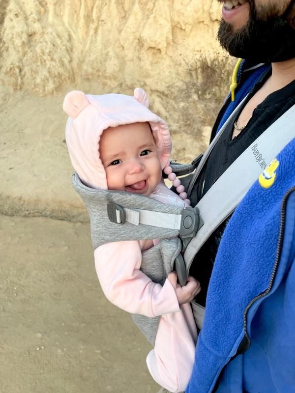 Baby in a pink hoodie and bear ears smiling in a gray carrier, worn by an adult in a blue jacket outdoors.