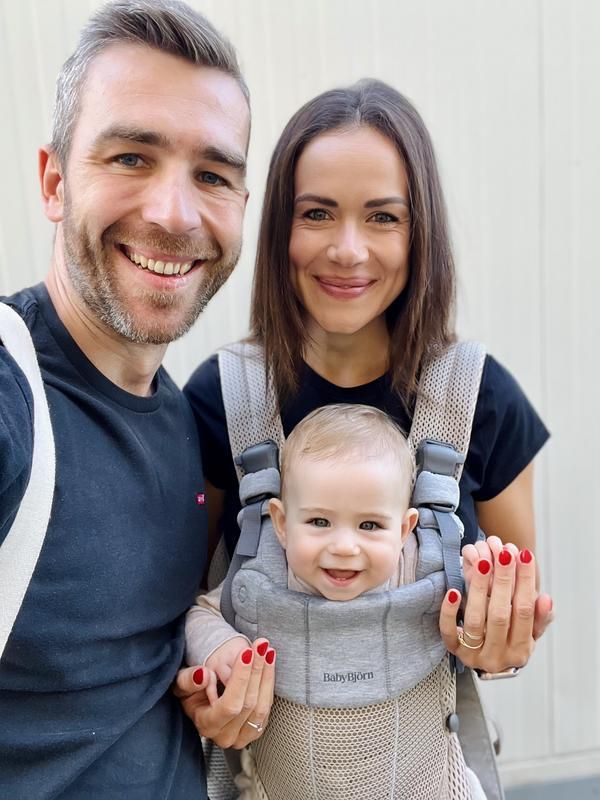 Smiling couple with a baby in a carrier. The baby is in front, supported by both parents, against a neutral background.