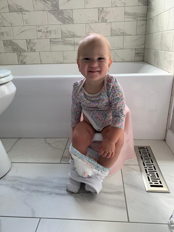 A smiling baby sits on a pink potty in a tiled bathroom, with a diaper around their ankles, in front of a bathtub.
