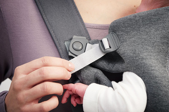 Close-up of a person adjusting a gray baby carrier strap. A baby's arm, wearing a white sleeve, is visible resting inside the carrier.