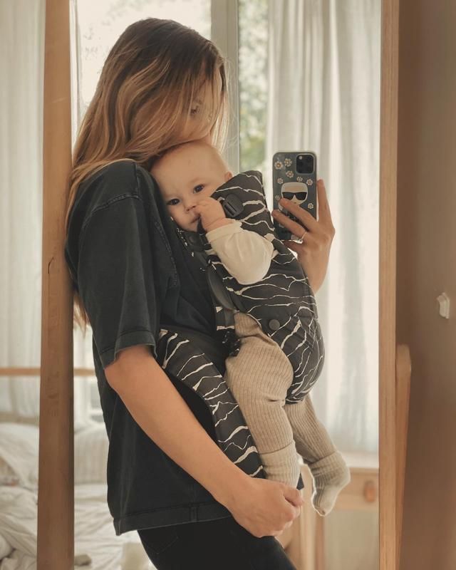 Woman taking a mirror selfie, holding a baby in a patterned carrier. Both face the camera, standing in a softly lit room.