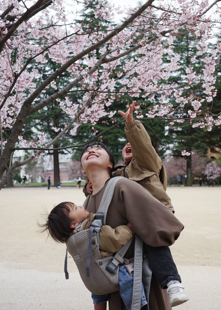 A woman with two children under blooming cherry blossoms; one child reaches up excitedly, while the other rests in a carrier.