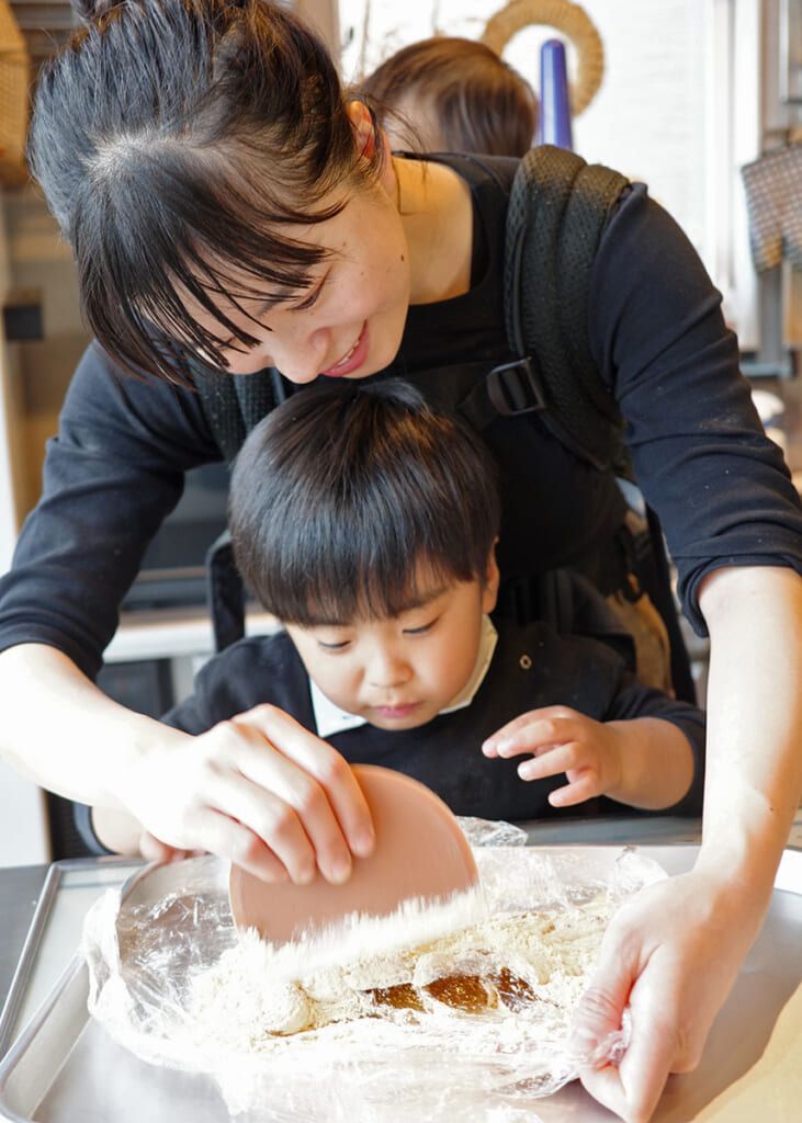 A woman helps a young child prepare dough on a tray. The child is using a scraper, and a baby is carried on the woman's back.