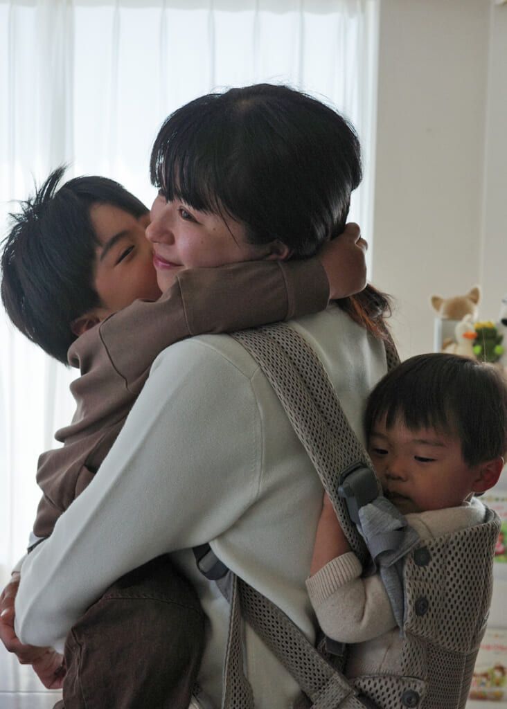 A woman smiles while holding a young child who hugs her. Another child in a carrier on her back. Bright window light in the background.
