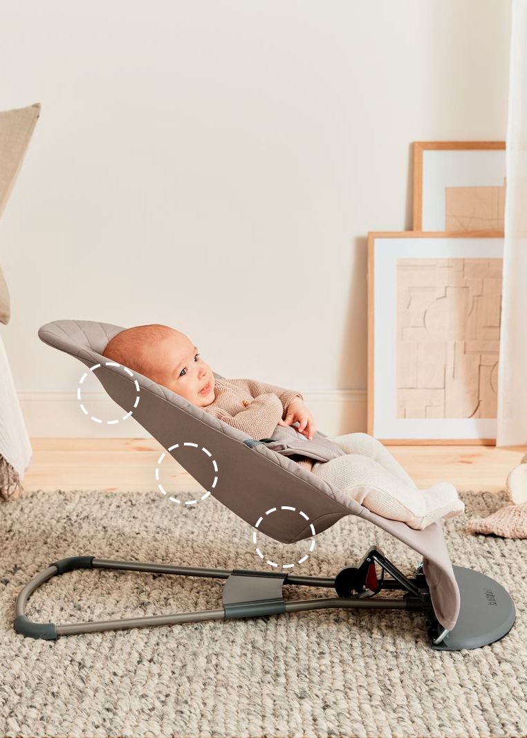 A baby in a beige outfit sits in a gray baby bouncer on a textured rug, with framed art and a curtain in the background.