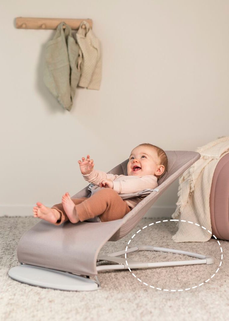 A baby smiling and reclining in a gray baby bouncer on a carpeted floor, with clothes hanging on a wall hook in the background.