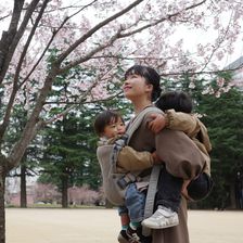 A woman smiles while carrying two young children under cherry blossoms in a park. The children are bundled in jackets, enjoying the serene setting.