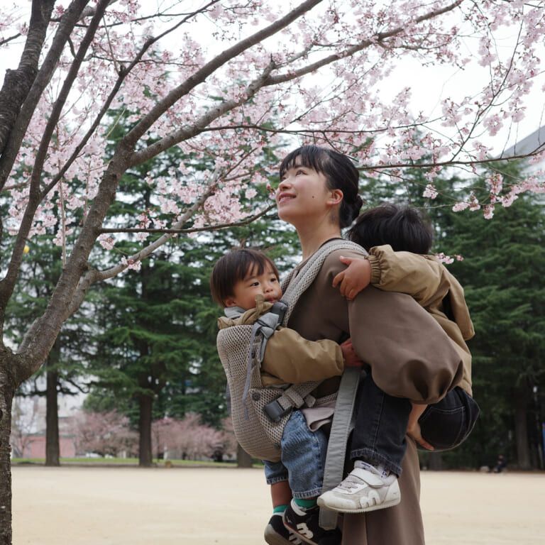 A woman smiles while carrying two young children under cherry blossoms in a park. The children are bundled in jackets, enjoying the serene setting.