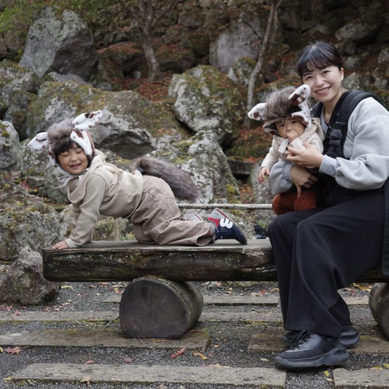 A smiling woman and two children wearing animal ear hats and tails pose on a wooden bench in a rocky outdoor setting.