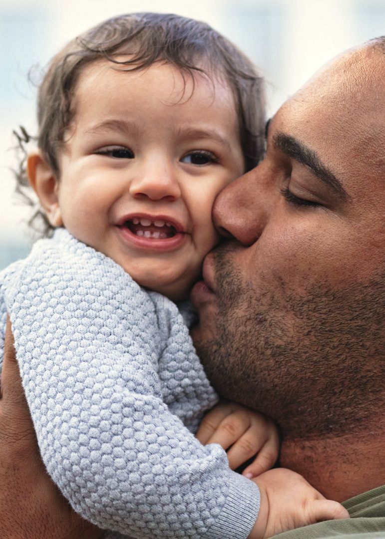 Adult kissing a smiling baby wearing a light gray sweater, while holding the baby close outdoors.