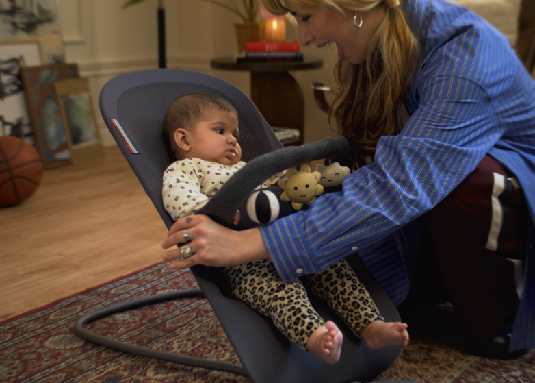 A woman in a blue striped shirt plays with a baby in a bouncer seat, surrounded by toys. A basketball and books are in the background.