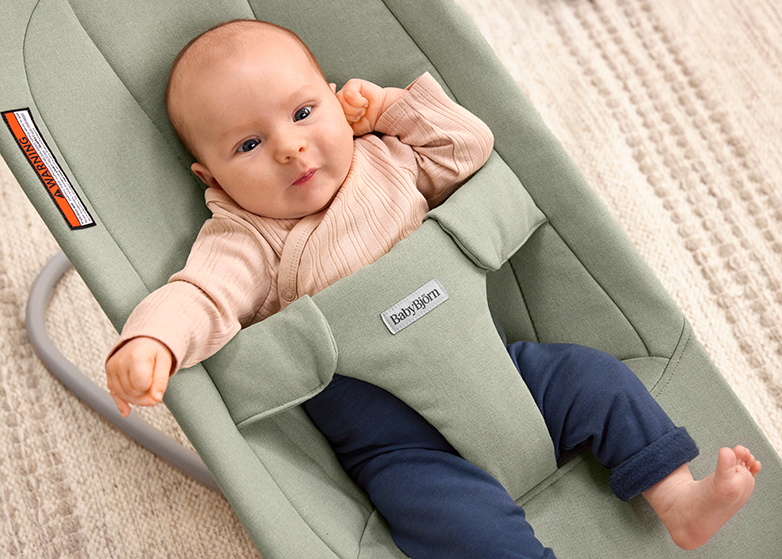 A baby in a light pink sweater and dark pants sits in a sage green BabyBjörn bouncer, looking up with one hand raised.
