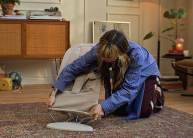 A person in a blue shirt kneeling on a patterned rug adjusts a beige baby bouncer in a cozy, warmly lit living room.