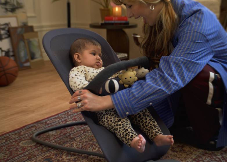 A woman smiles at a baby sitting in a bouncer with hanging toys, in a cozy room with a patterned rug and a basketball in the background.
