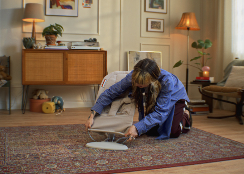 A person is kneeling on a rug in a cozy living room, adjusting a beige baby bouncer. The room has a wooden cabinet and potted plants.