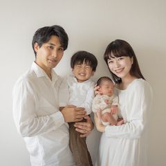 A smiling family of four poses together against a plain background. The parents hold a young child and a newborn baby, all dressed in light-colored clothing.