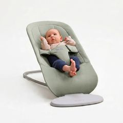A baby sits comfortably in a light green baby bouncer against a plain background, wearing a light sweater and dark pants.