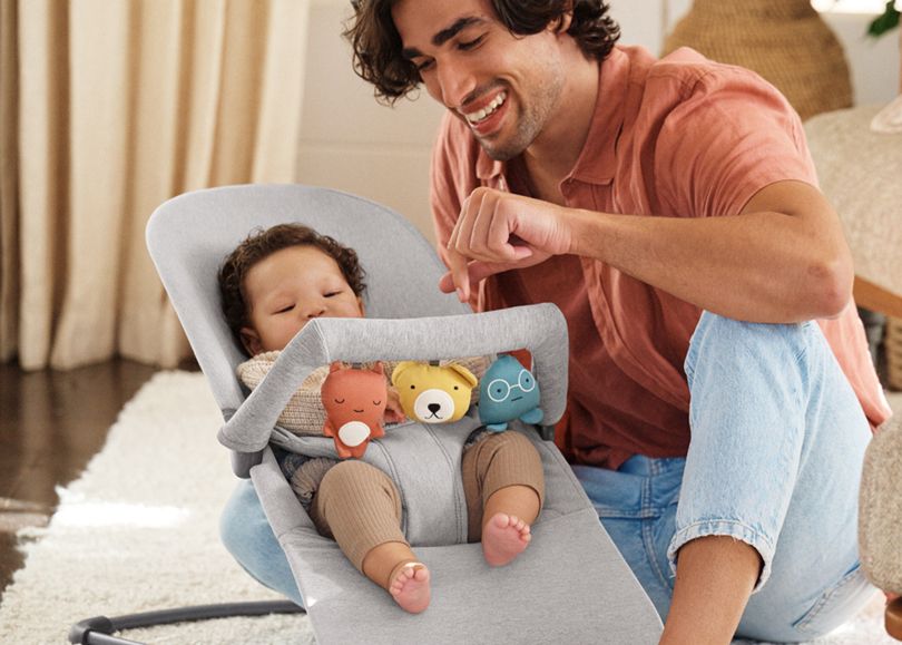 A man smiles at a baby in a bouncer with soft toy bar attached. The baby is relaxed, wearing a beige outfit.