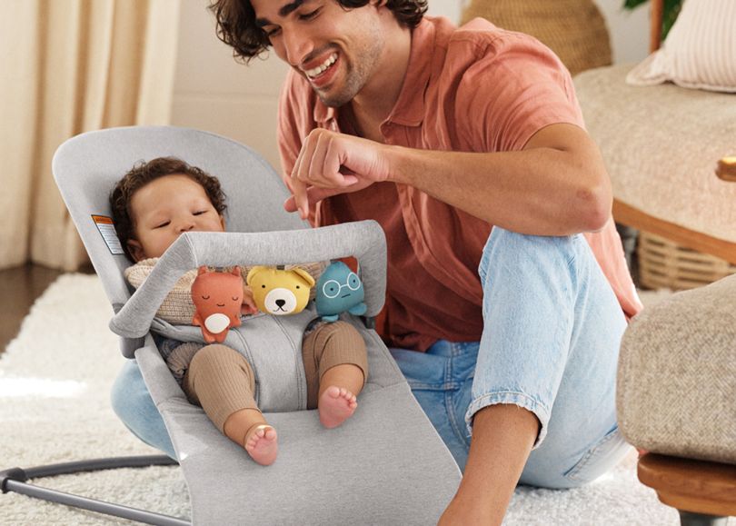 A smiling man interacts with a baby in a gray bouncer from BabyBjörn and toy bar Soft Friends.
