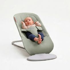 A baby is lying in a light green baby bouncer, looking up with arms and legs raised. The background is a neutral, soft white.