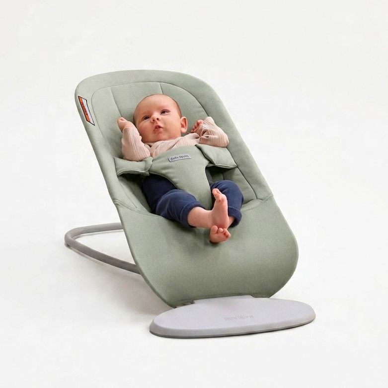 A baby is lying in a light green baby bouncer, looking up with arms and legs raised. The background is a neutral, soft white.