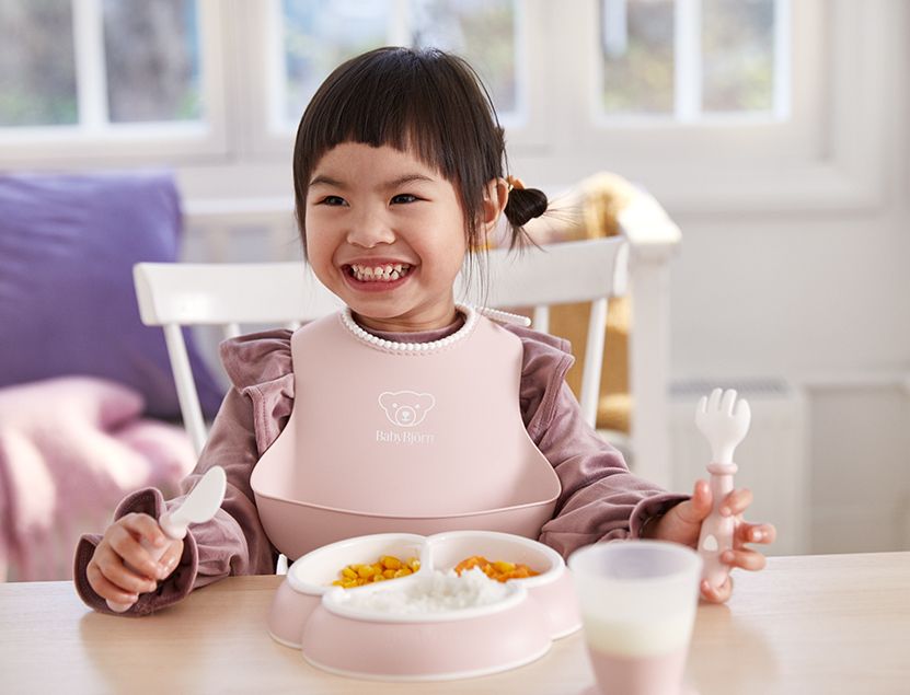 Smiling toddler in a pink bib sits at a table with a partitioned plate of food and a cup, holding a spoon and fork in a bright room.