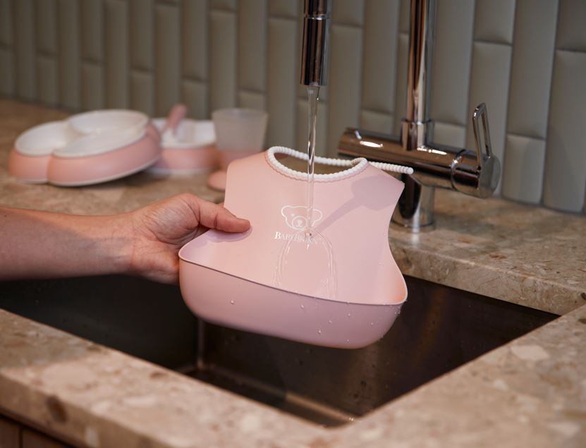 A person rinses a pink silicone bib under a running faucet in a kitchen sink, with more baby items in the background.