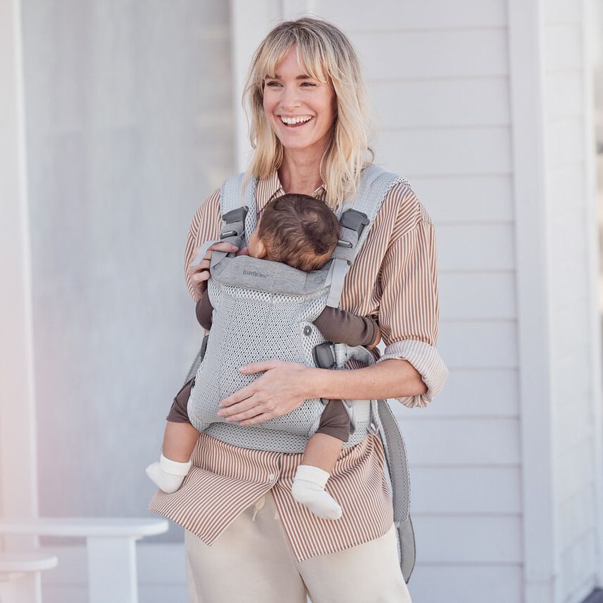 Smiling woman carrying a baby in a gray baby carrier, standing outside a white building.