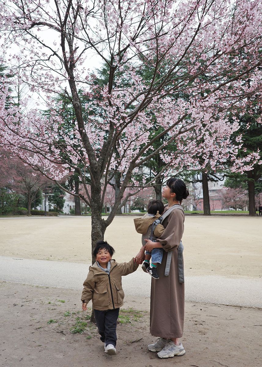 A woman with two children stands under a blooming cherry blossom tree in a park. The older child smiles and holds her hand.