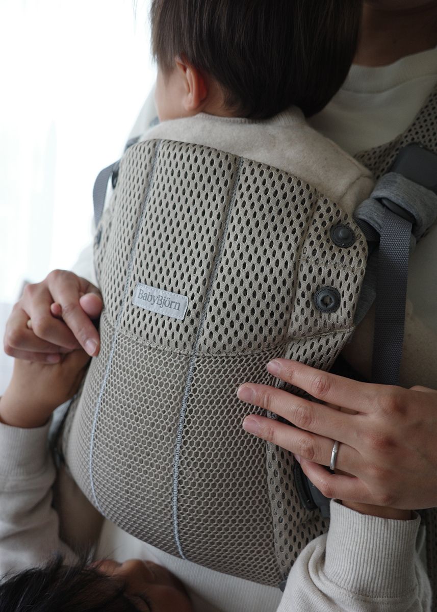 Baby in a gray mesh carrier held by an adult, with hands supporting the back. The carrier has a "BabyBjörn" label.