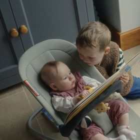 A child sits by a baby in a bouncy seat, both engaged with a picture book. The baby is holding a toy, and they are near a cabinet.