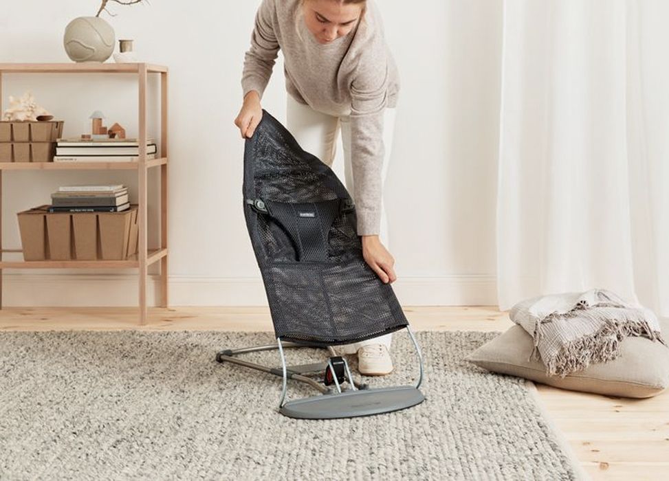 Person assembling a black mesh baby bouncer on a gray rug in a cozy room with a wooden shelf and cushions in the background.