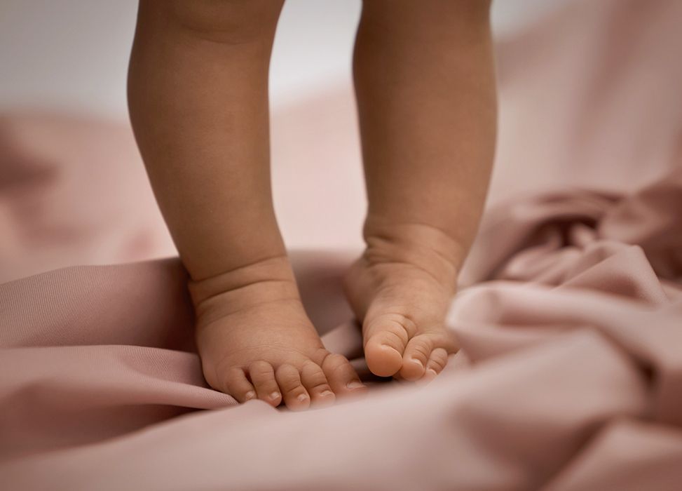 Close-up of a baby's feet standing on soft, pink fabric, with focus on tiny toes and smooth skin.