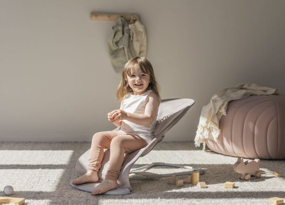 A smiling child sits in a soft chair on a sunlit carpeted floor, surrounded by wooden toys, with a pouf and hanging clothes in the background.