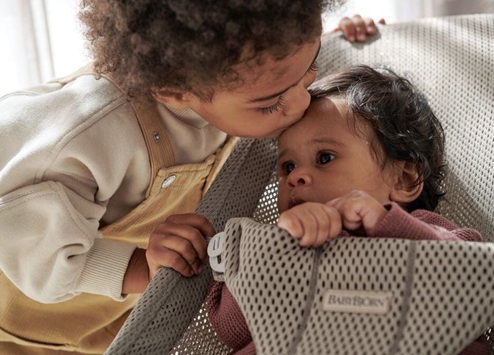 A young child gently kisses a baby on the forehead as the baby lies in a mesh baby carrier.