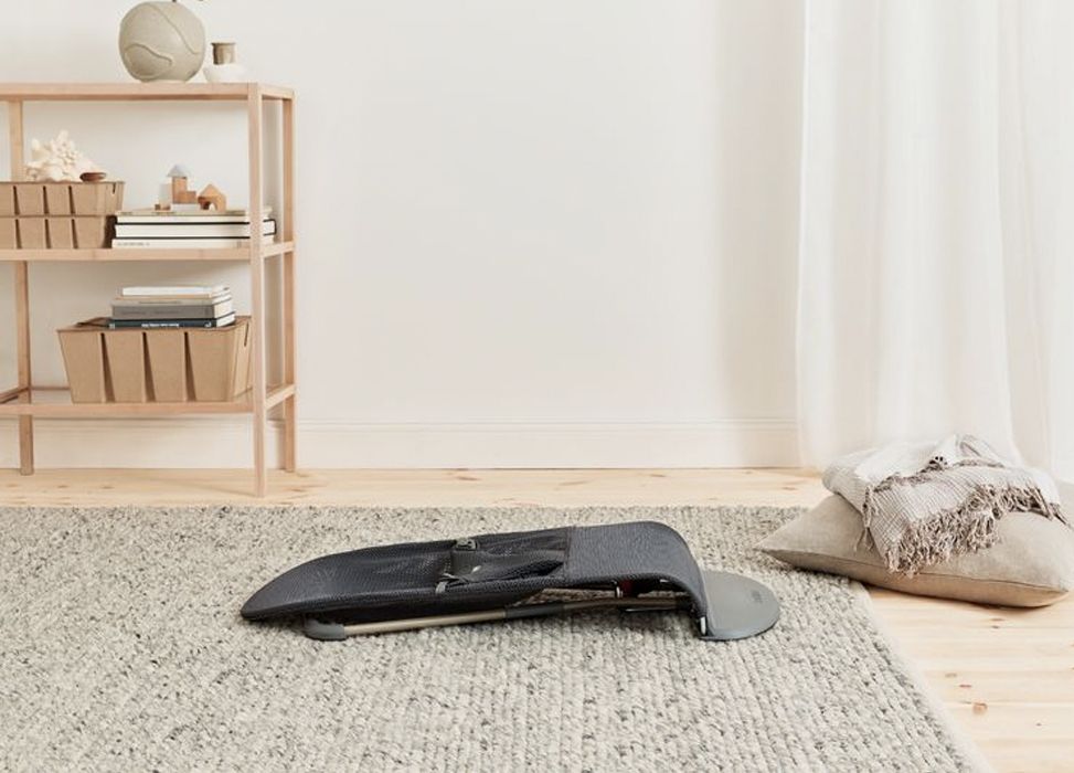 A baby bouncer on a carpeted floor near a wooden shelf with baskets and a pillow, in a bright, minimalist room.