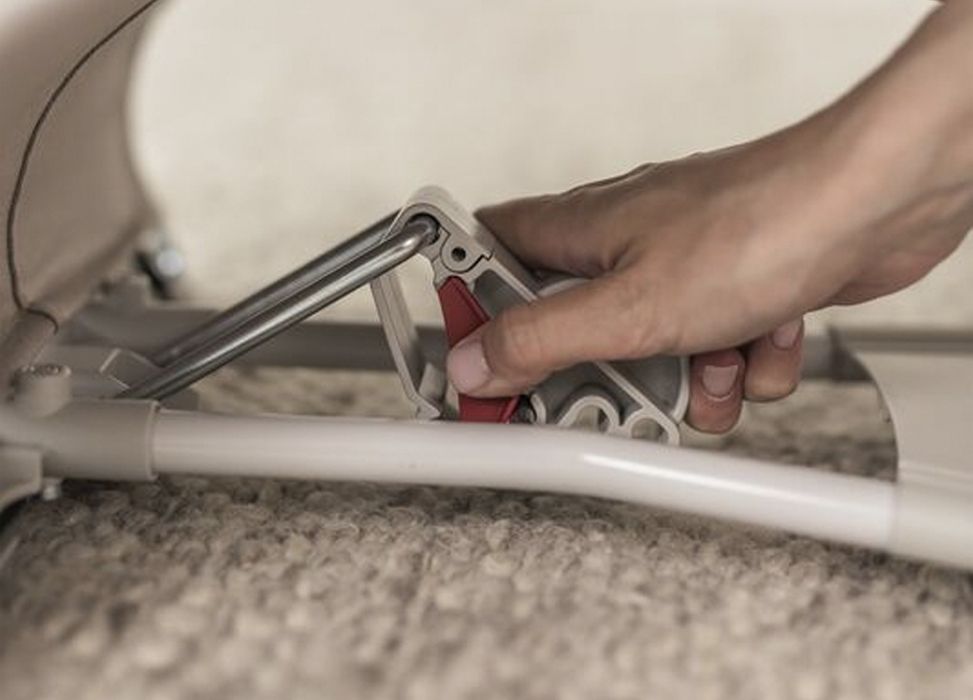 Hand adjusting a red lever on a folding mechanism of a beige stroller, placed on a carpeted floor.